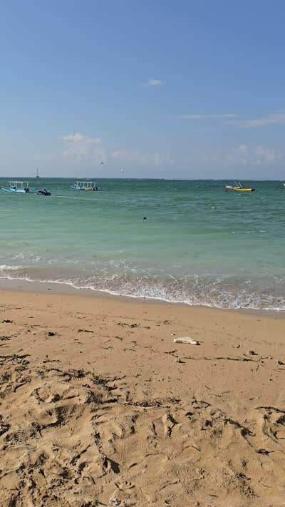 Vertical video of Mertasari Beach in Bali, showing golden sand, turquoise ocean with boats, and a clear blue sky. - stock photo