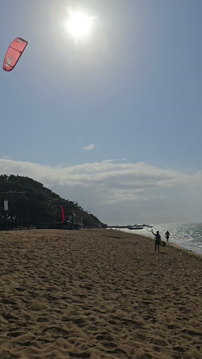 Vertical footage of Pantai Bopel beach in Bali with a red kite, ocean waves, and people walking on sand under a sunny sky. - stock photo