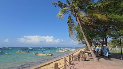 Tracking shot of Pantai Duyung beach in Bali with traditional Jukung boats, palm trees, and clear turquoise water under a blue sky. - stock photo
