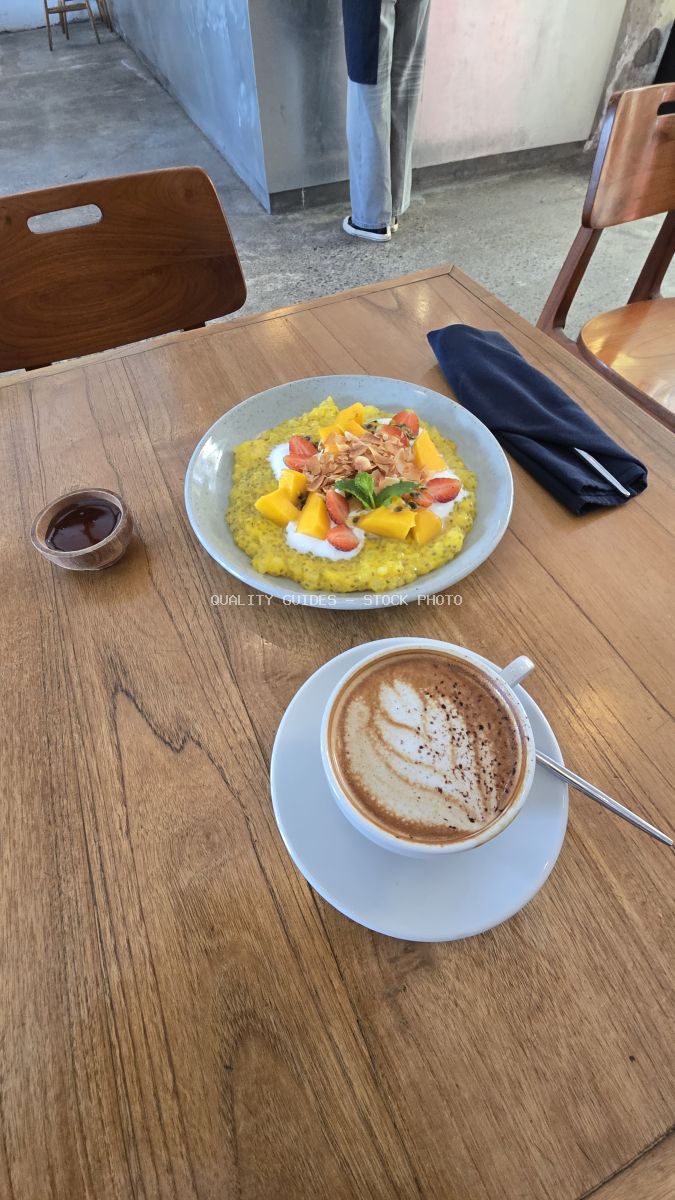 Overhead view of a colorful tropical breakfast bowl with mango and strawberries, and a latte on a wooden cafe table. - stock photo