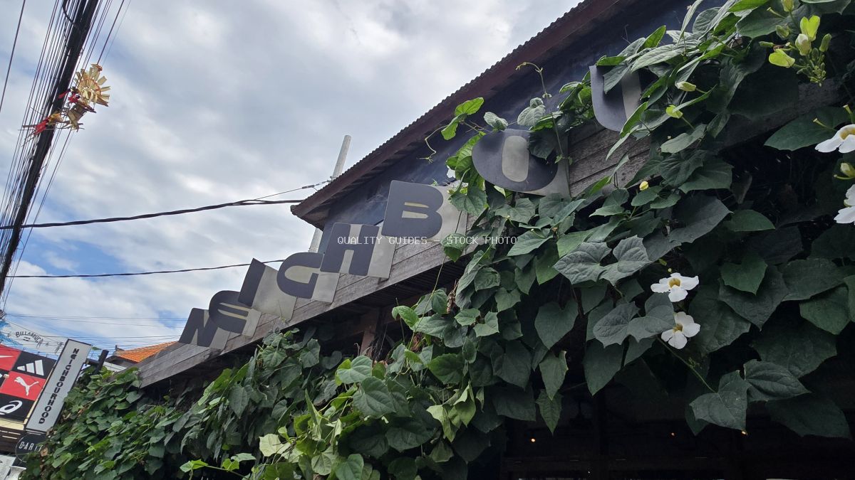 Exterior of Neighbourhood Food cafe in Bali, covered in green vines and white flowers, with a visible sign and power lines. - stock photo