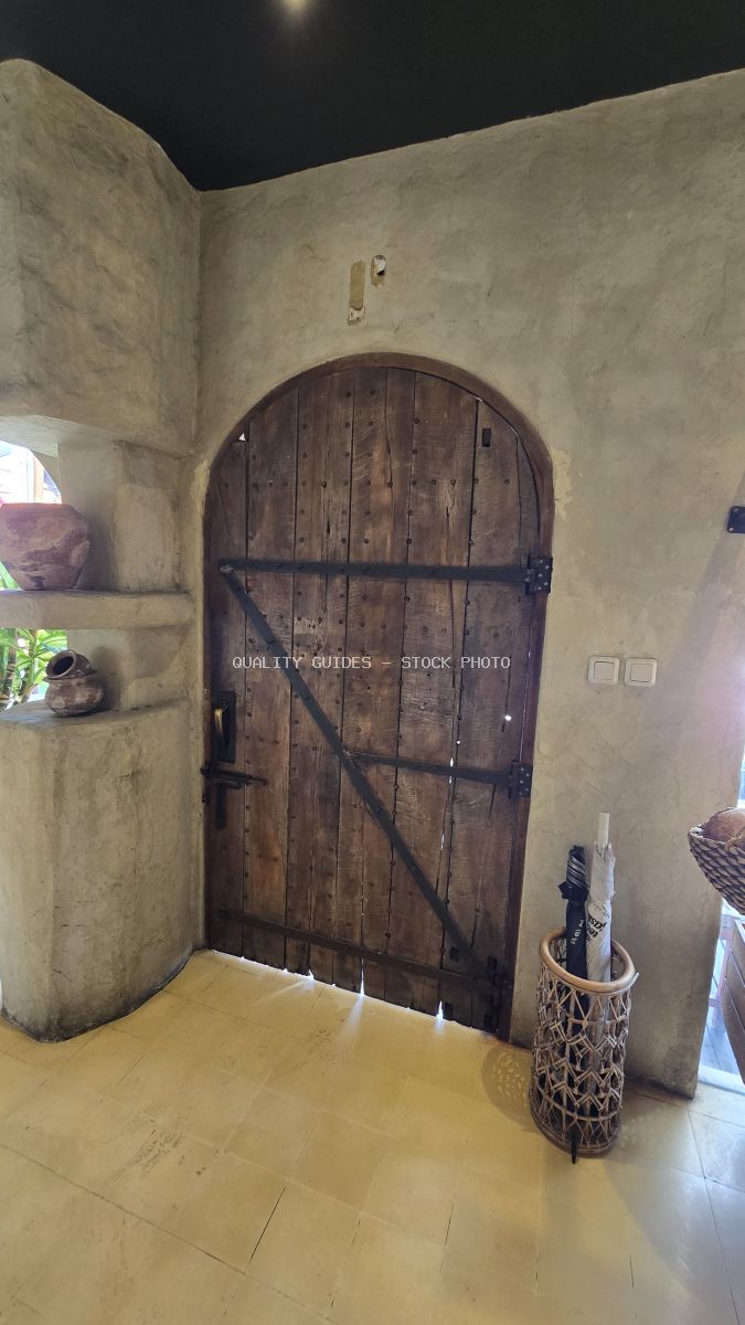 Arched rustic wooden door with metal straps in a textured stucco wall, flanked by clay pots and an umbrella stand. - stock photo