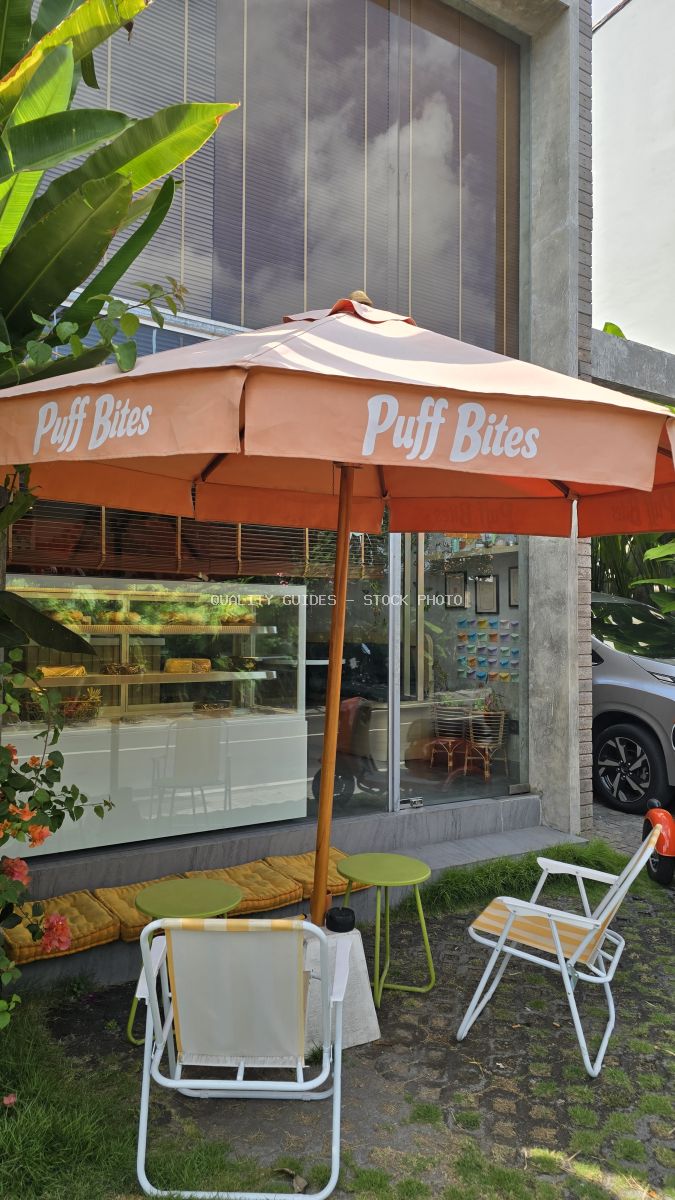 Exterior of Puff Bites cafe in Bali with an orange umbrella, outdoor chairs, and a pastry display visible through the window. - stock photo
