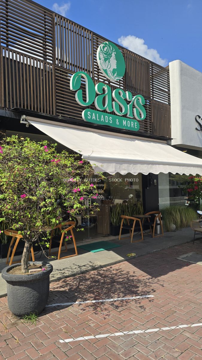 Exterior of Oasis Salads & More restaurant in Bali with green sign, white awning, outdoor seating, and potted pink flowers. - stock photo