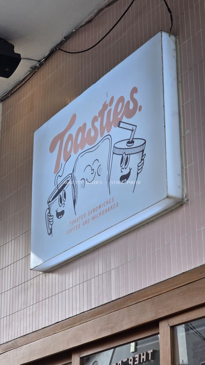 Illuminated sign for 'Toasties' cafe with cartoon toast, coffee, and milkshake characters on a tiled building facade. - stock photo