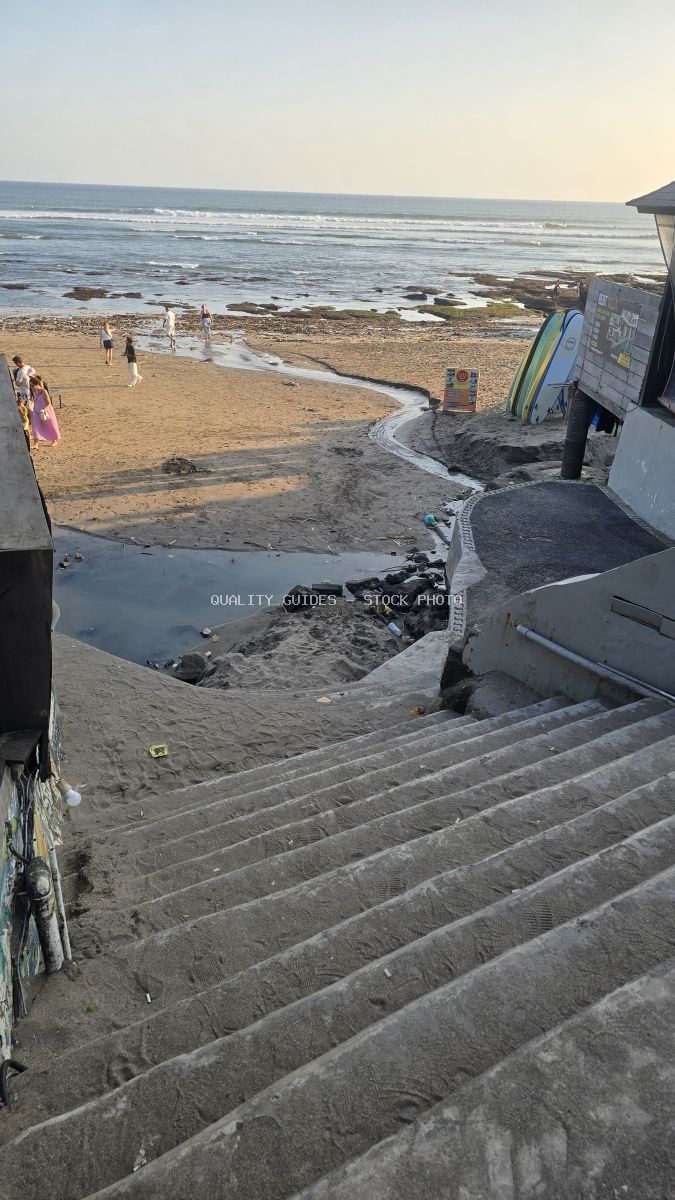 Concrete stairs descending to a sandy beach with a stream, ocean, and distant people under a soft sky. - stock photo