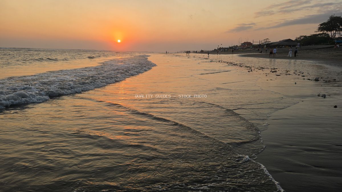 Sunset over Nelayan Beach in Bali with golden sky, ocean waves, and distant people. - stock photo
