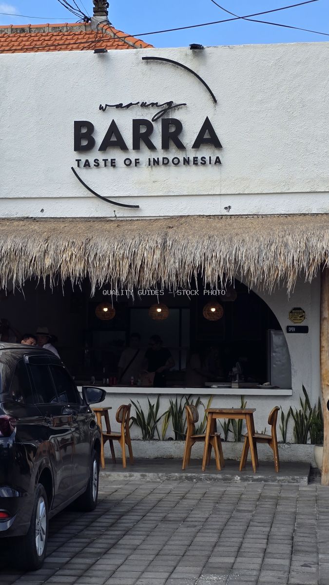 Exterior view of Warung Barra restaurant in Bali, featuring a white building, thatched roof, outdoor wooden seating, and a parked black car. - stock photo
