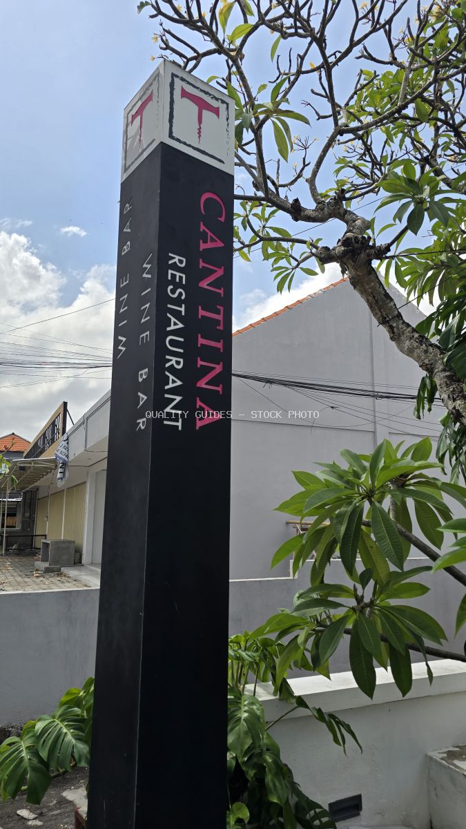 Black signpost for Cantina Restaurant Wine Bar in Bali, with green trees and blue sky. - stock photo
