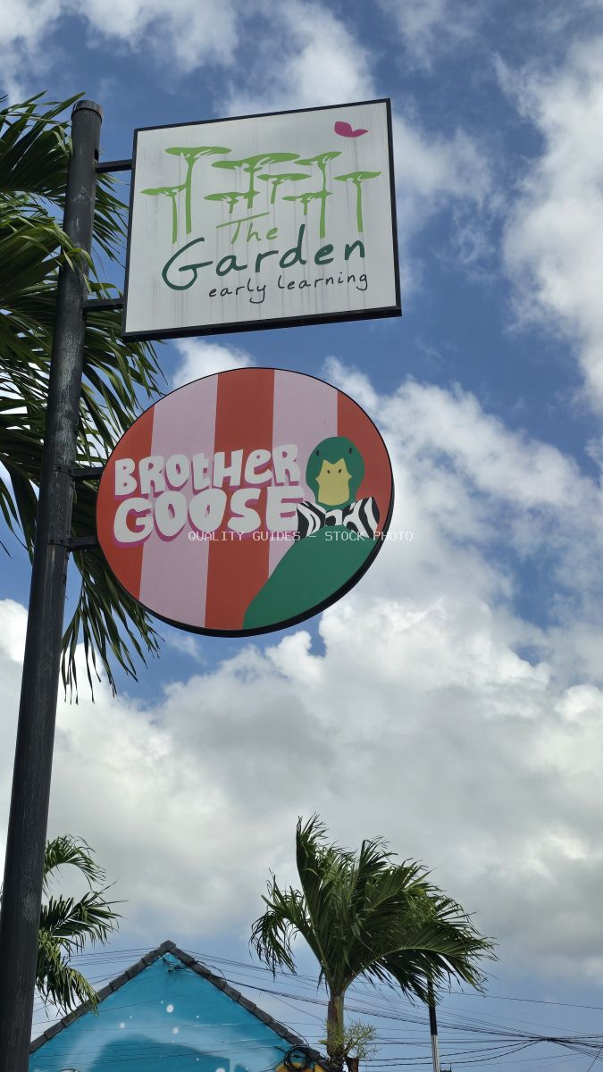 Signs for Brother Goose Cafe and The Garden Early Learning against a blue sky with clouds and palm trees in Bali. - stock photo