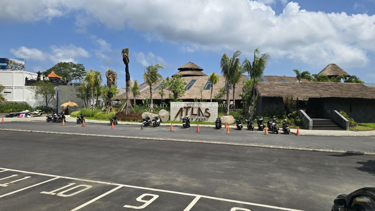 Exterior view of Atlas Beach Fest in Bali, featuring a large thatched-roof building, palm trees, and a parking lot with motorbikes under a sunny sky. - stock photo