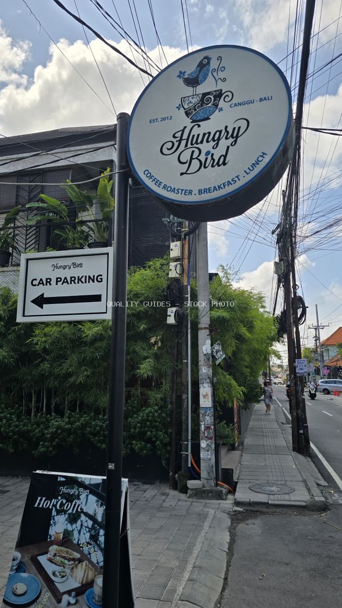 Exterior view of Hungry Bird Coffee Roaster sign in Canggu, Bali, with street, parking sign, and tropical foliage. - stock photo