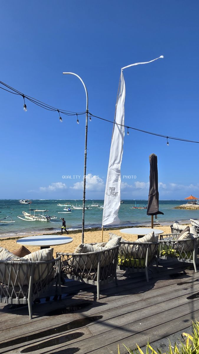Outdoor lounge at Pier Eight Bali beach club with ocean view, traditional boats, and a person walking on the sand. - stock photo