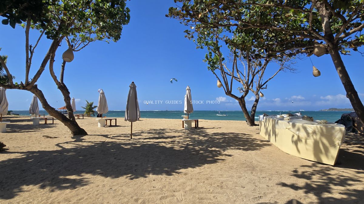Mertasari Beach in Bali with sand, ocean, tropical trees, closed umbrellas, and distant kitesurfers under a clear blue sky. - stock photo