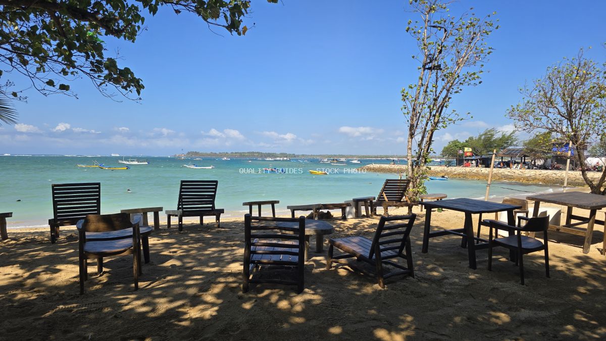 Empty wooden chairs and tables on the sandy Mertasari Beach in Bali, with turquoise ocean and traditional boats under a clear blue sky. - stock photo