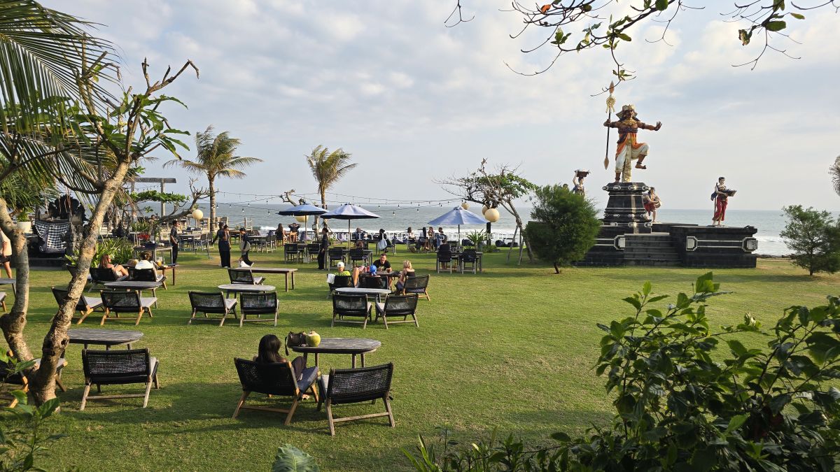 People relaxing at an outdoor beach club on a green lawn overlooking the ocean in Bali, with a large Balinese statue. - stock photo