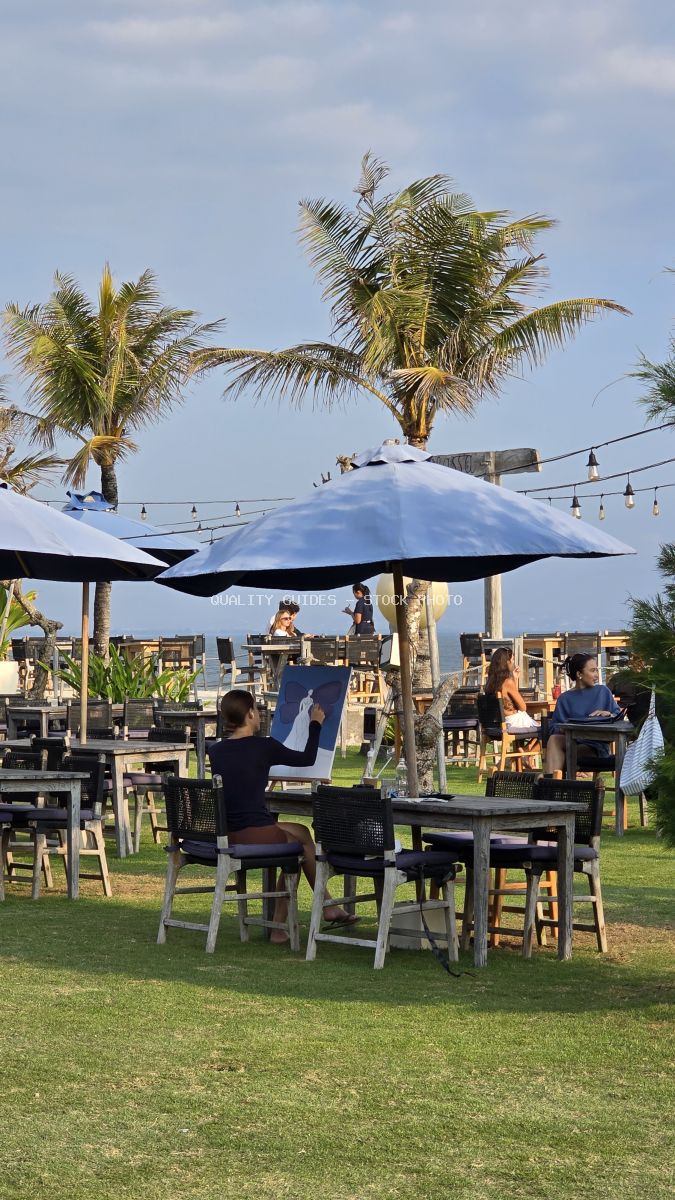 Person painting at an outdoor beach club in Bali with palm trees and blue umbrellas. - stock photo