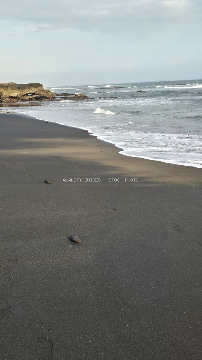 Seseh Beach in Bali with dark volcanic sand, ocean waves, and rocky coastline under an overcast sky. - stock photo