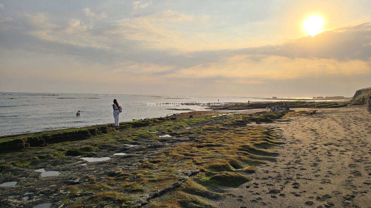 Sunset at Cemagi Beach, Bali, with a person on mossy rocks looking at the ocean and distant groups. - stock photo