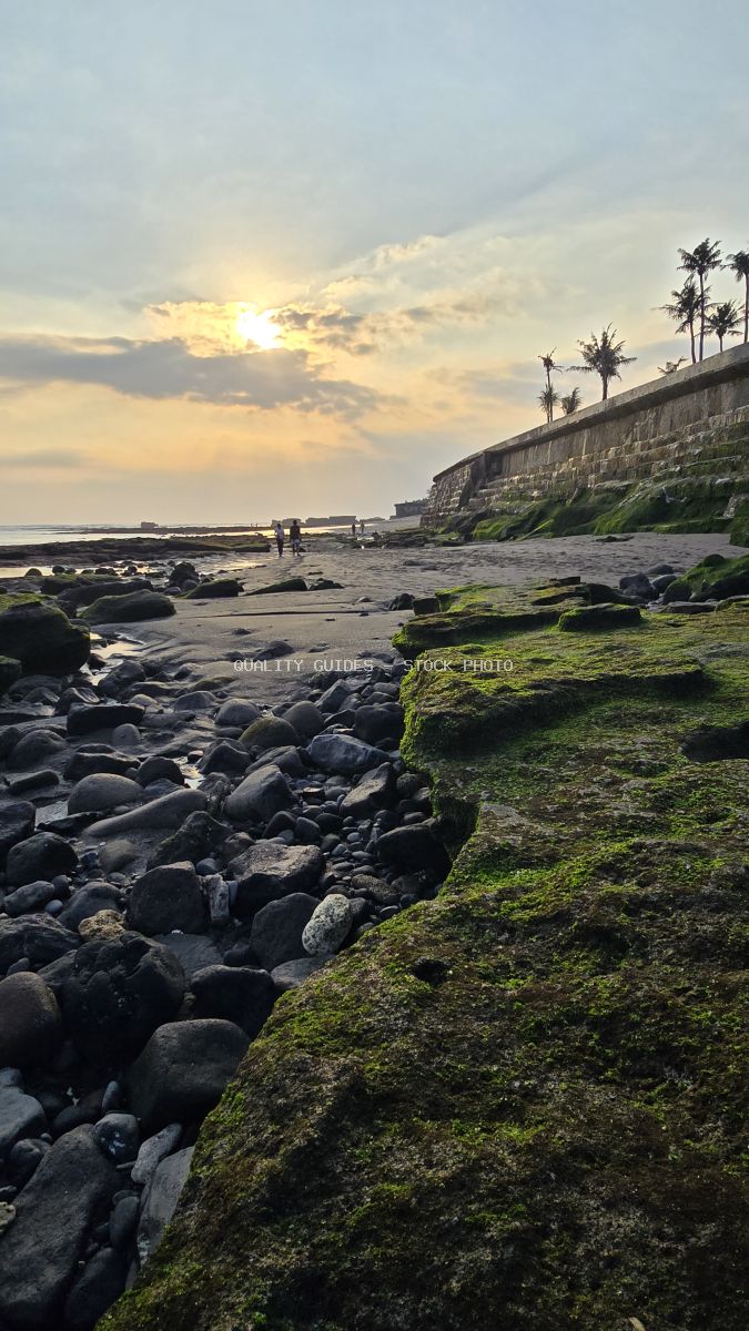 Sunset at Cemagi Beach, Bali, with mossy rocks, dark sand, distant figures, and palm trees. - stock photo