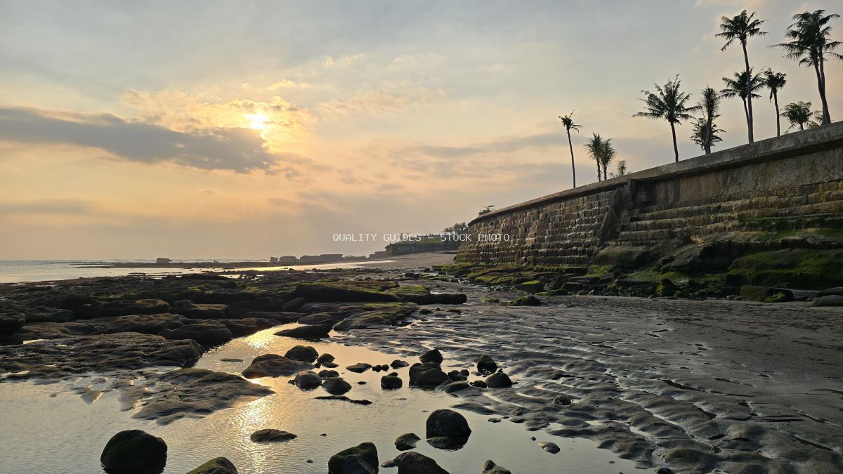 Sunset over Cemagi Beach, Bali, with rocky shoreline, tide pools, and palm trees on a retaining wall. - stock photo