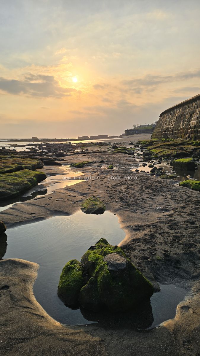 Sunset over a rocky beach in Bali with tide pools, mossy rocks, and a stone wall. - stock photo