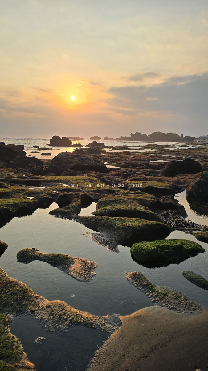 Sunset over a rocky beach with tide pools reflecting the golden sky at Mengening Beach, Bali. - stock photo