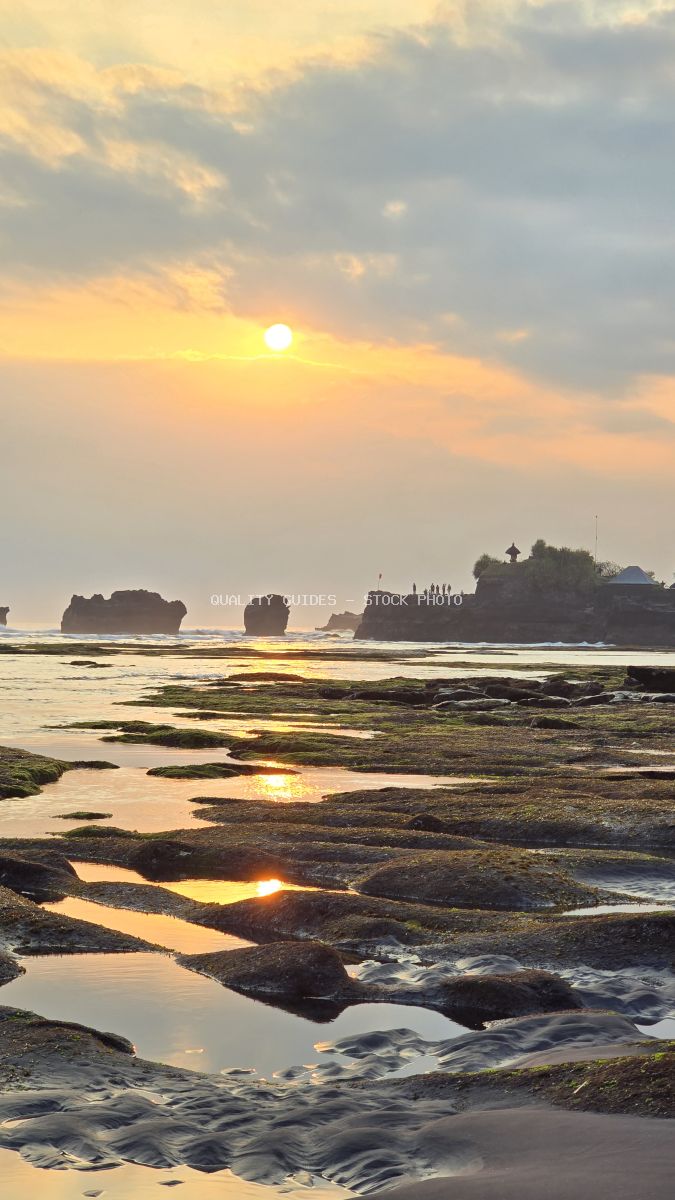 Sunset over Tanah Lot Temple in Bali, with golden light reflecting in tidal pools on a rocky beach. - stock photo