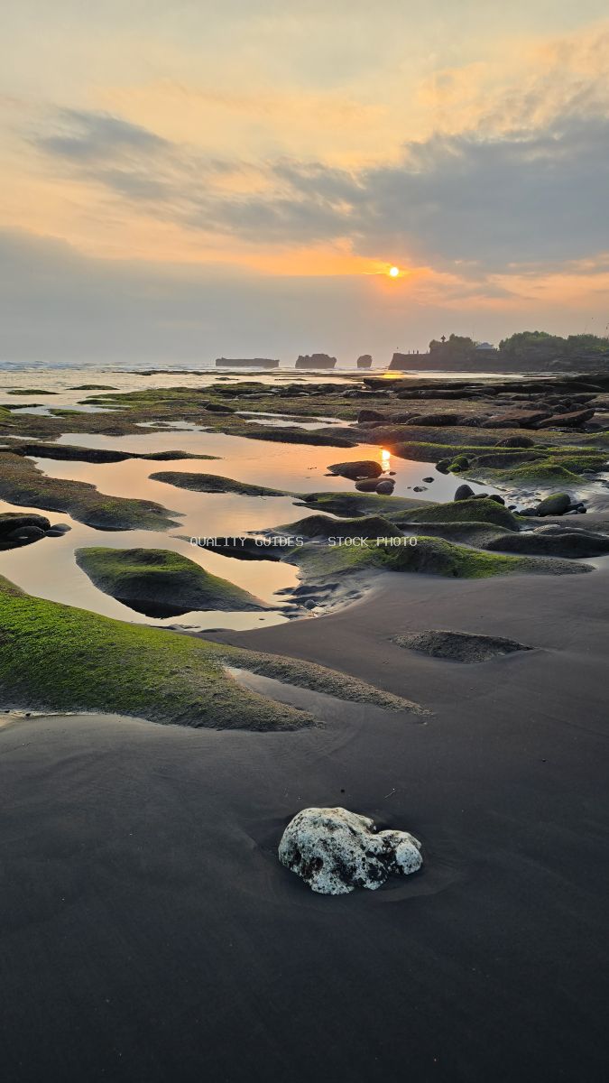 Sunset over Mengening Beach, Bali, with black sand, green mossy rocks, and tidal pools reflecting the golden sky. - stock photo
