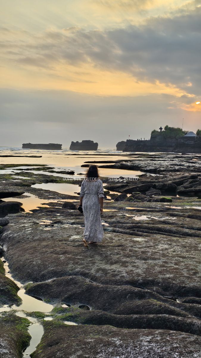 Woman in a dress walking on a rocky beach at sunset with Tanah Lot temple in the distance. - stock photo