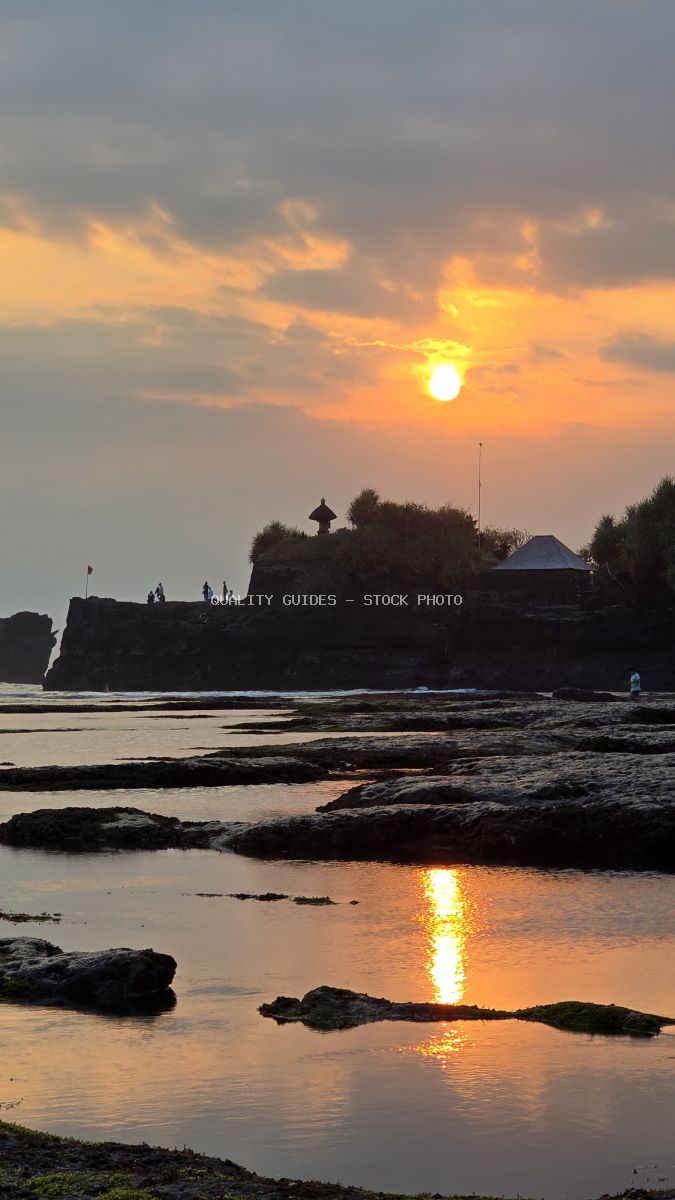 Sunset over a Balinese temple on a rocky beach with golden reflections on water. - stock photo