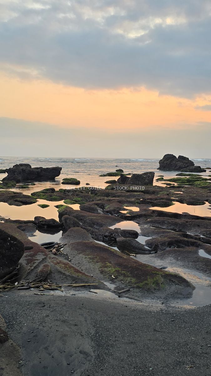 Rocky beach at sunset with volcanic rocks, tidal pools, and ocean waves under an orange and grey sky in Bali. - stock photo