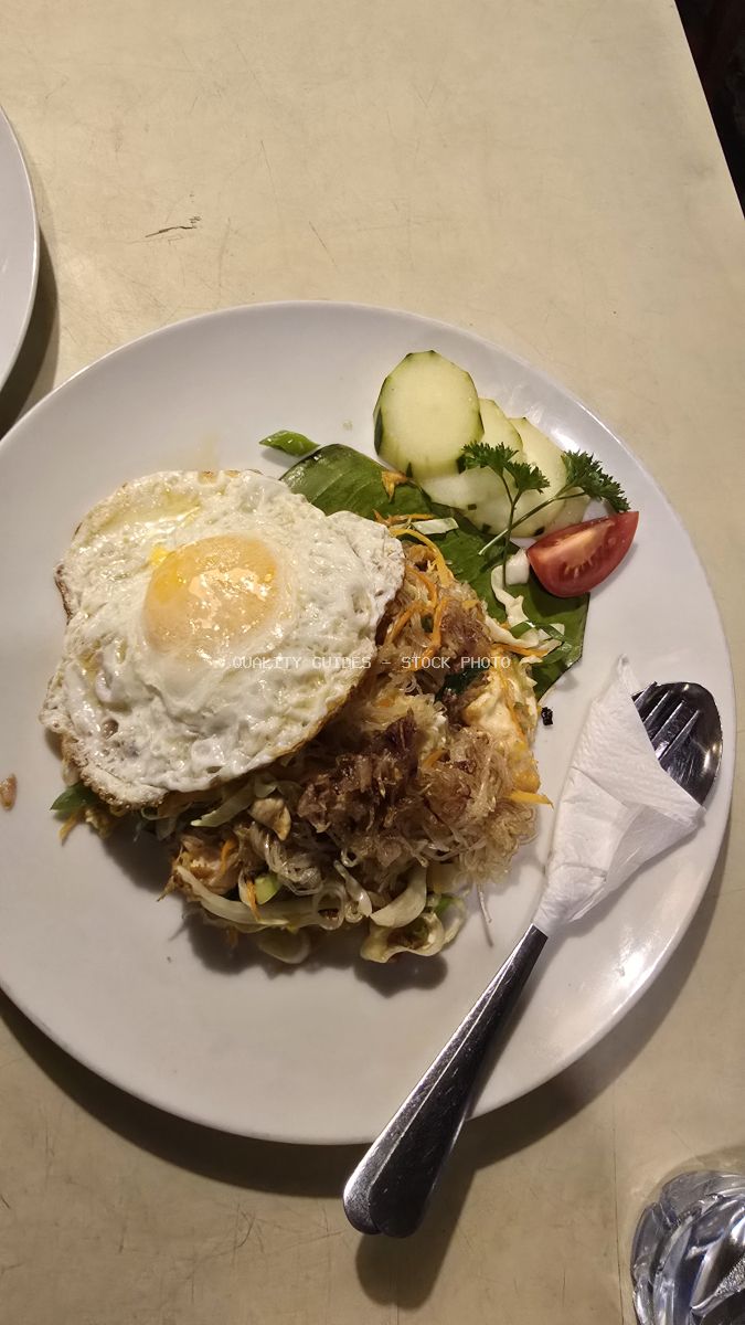 Plate of Indonesian fried noodles with a fried egg, cucumber, and tomato on a white plate. - stock photo