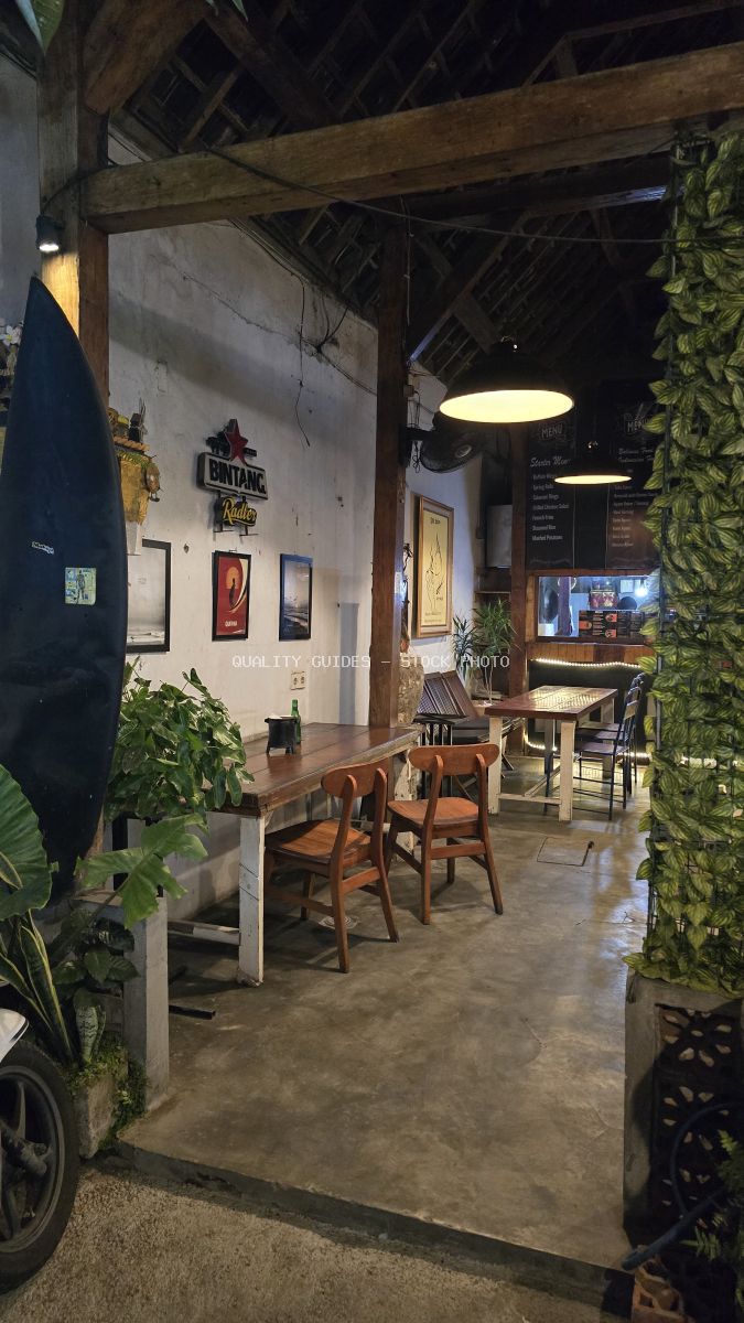 Interior of a rustic Balinese cafe with wooden tables, chairs, exposed beams, a Bintang sign, and tropical plants, illuminated by warm pendant lights. - stock photo