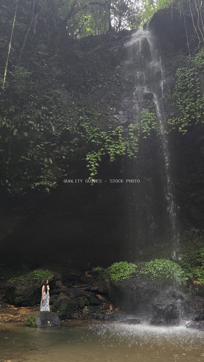 Woman in white dress standing at base of tall waterfall surrounded by tropical jungle vegetation in Bali - stock photo