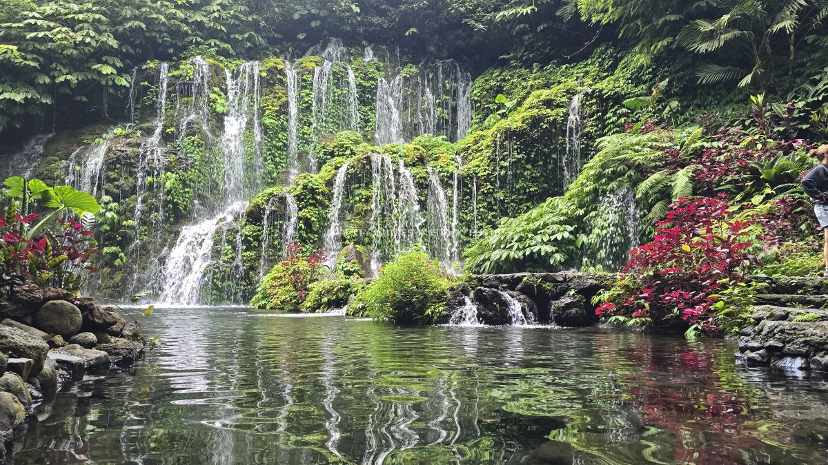 Multi-tiered waterfall flowing into emerald pool surrounded by lush tropical jungle and red flowering plants in Bali - stock photo