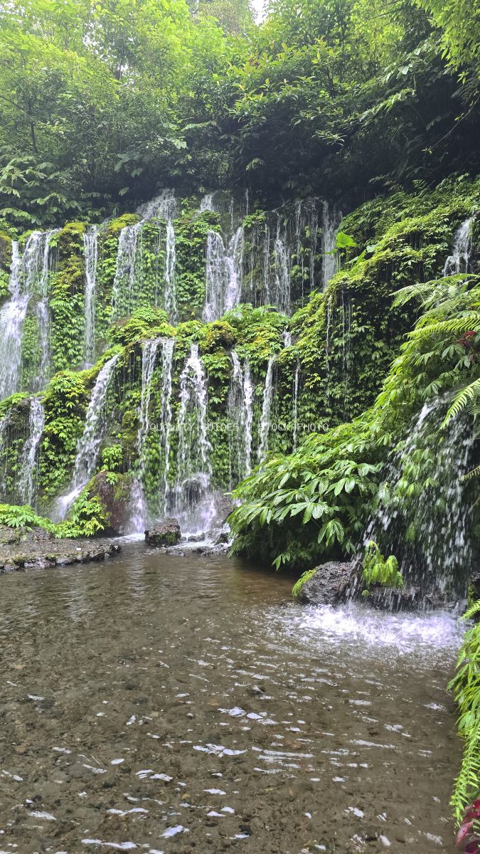 Multi-tiered waterfall cascading down moss-covered cliff surrounded by lush tropical vegetation in Bali - stock photo