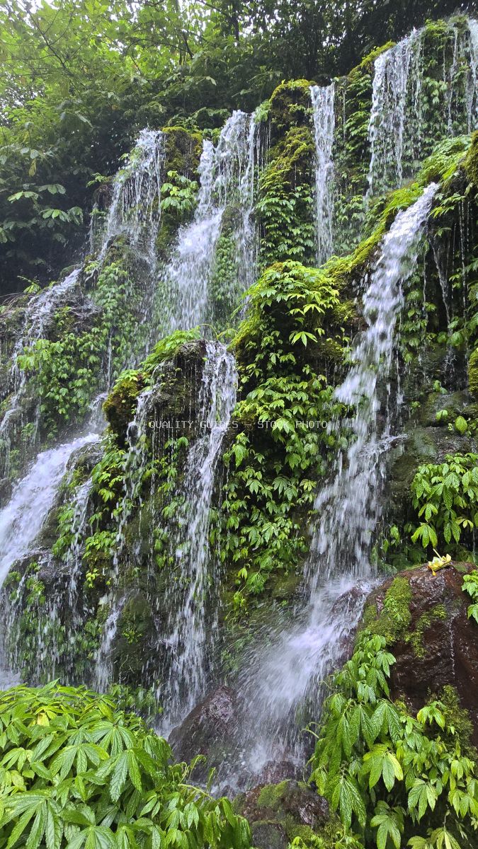 Multi-tiered waterfall with white cascading water surrounded by lush green tropical vegetation and moss-covered rocks in Bali - stock photo