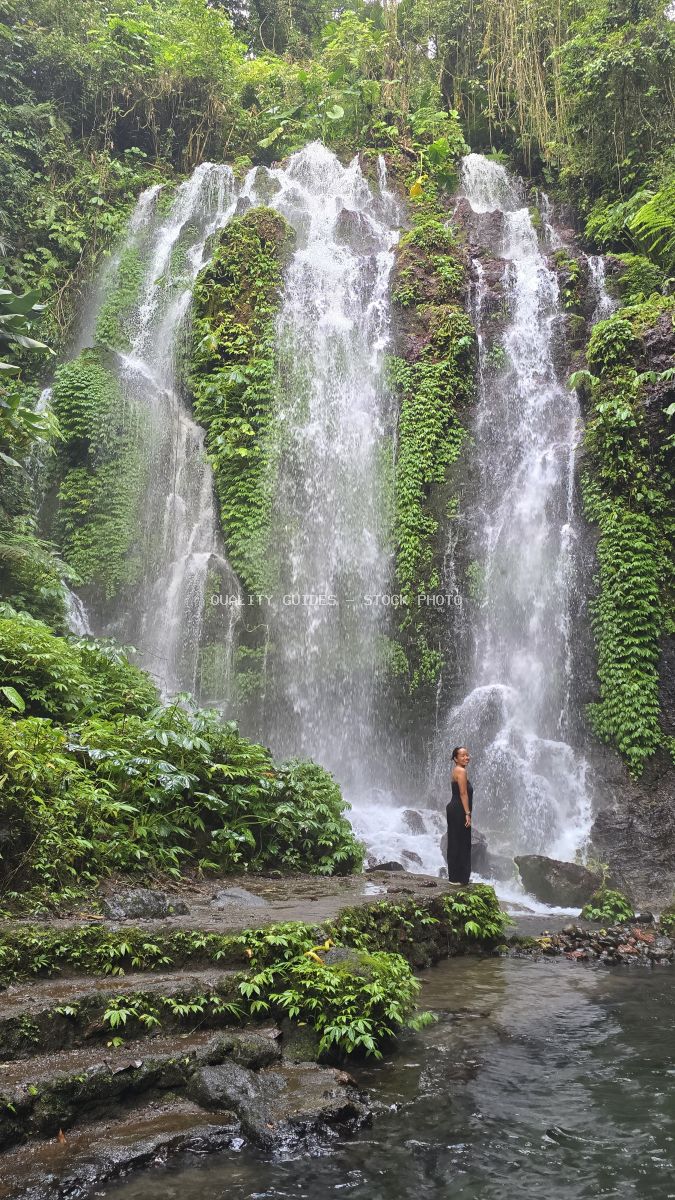 Person standing at base of multi-tiered waterfall surrounded by lush green jungle vegetation in Bali - stock photo