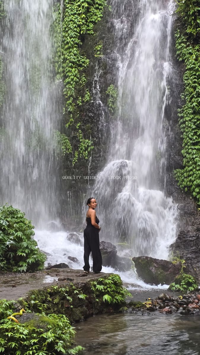 Woman standing at base of tall waterfall with ivy-covered cliffs in tropical Bali jungle - stock photo