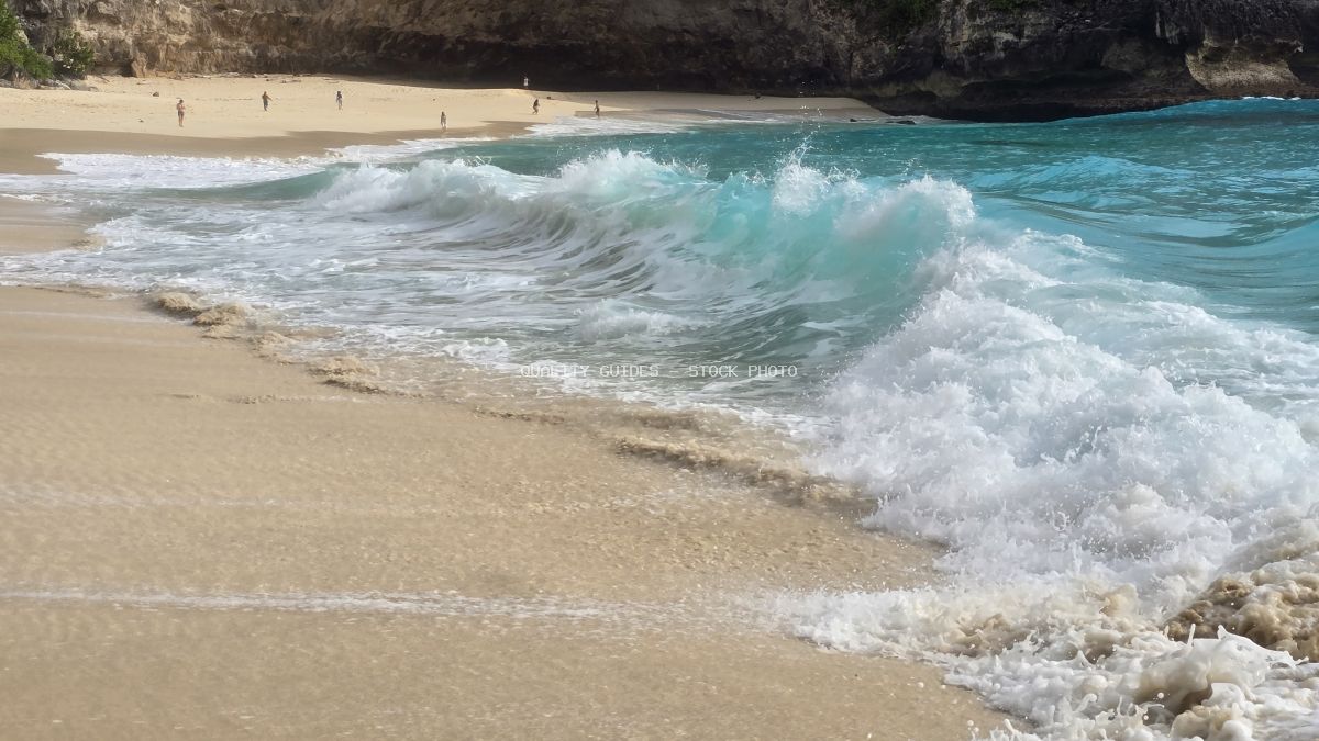 Turquoise waves crashing on sandy Kelingking Beach in Bali with limestone cliffs and beachgoers - stock photo