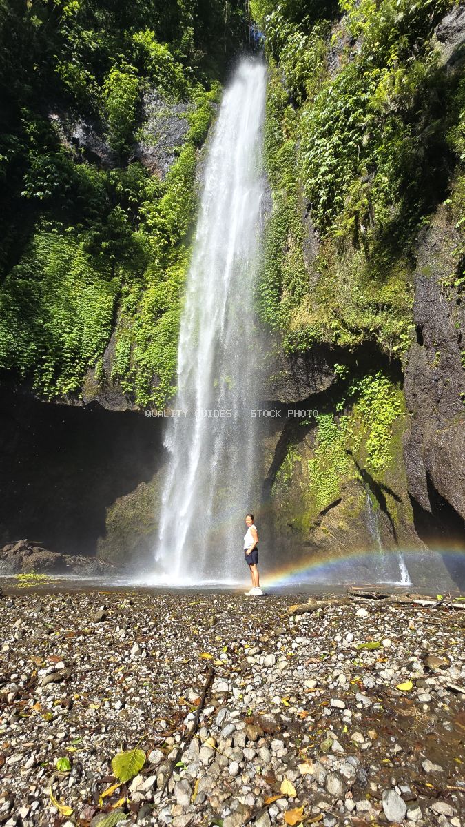 Traveler standing at base of Pucak Manik Waterfall in Bali with rainbow in mist, surrounded by lush green vegetation - stock photo