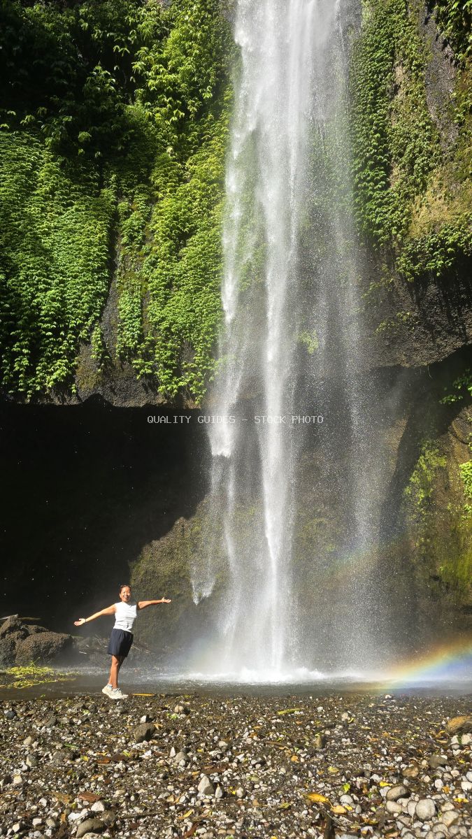 Woman with arms outstretched at Pucak Manik Waterfall in Bali with rainbow in mist - stock photo