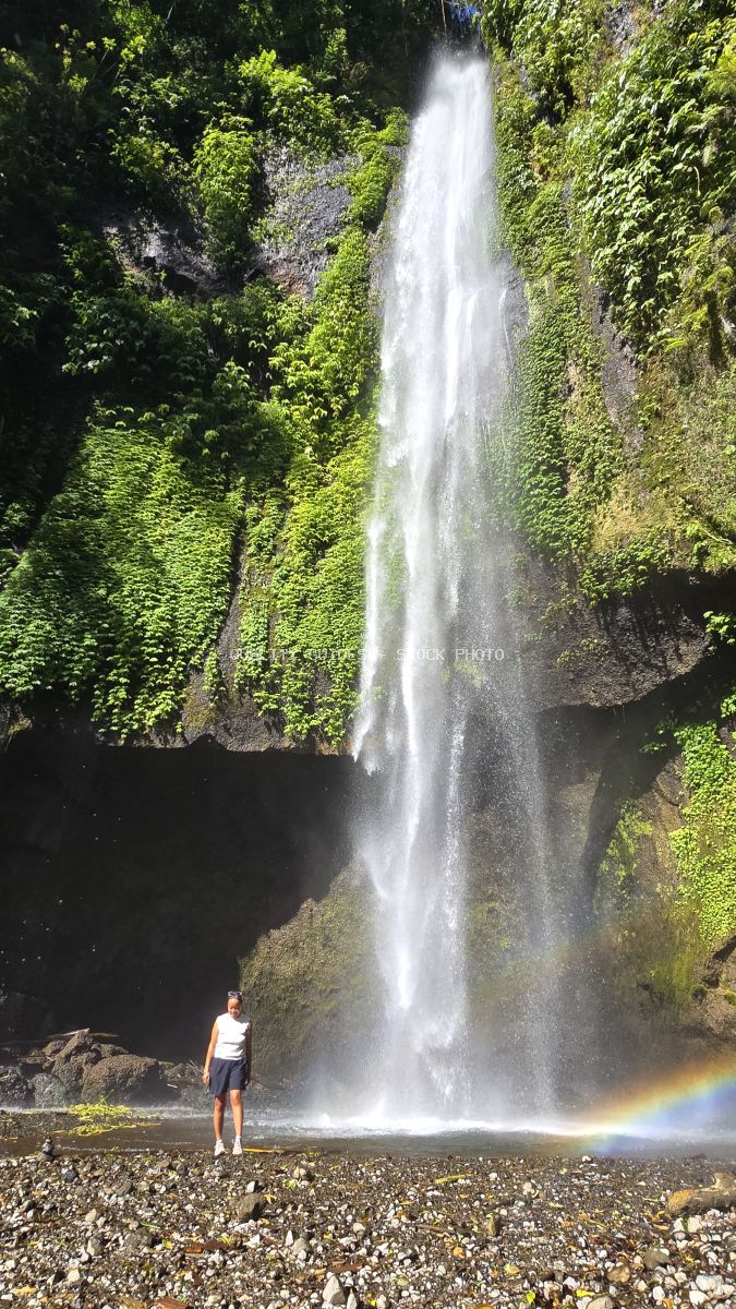 Solo traveler standing before a tall waterfall with rainbow mist in Bali jungle - stock photo