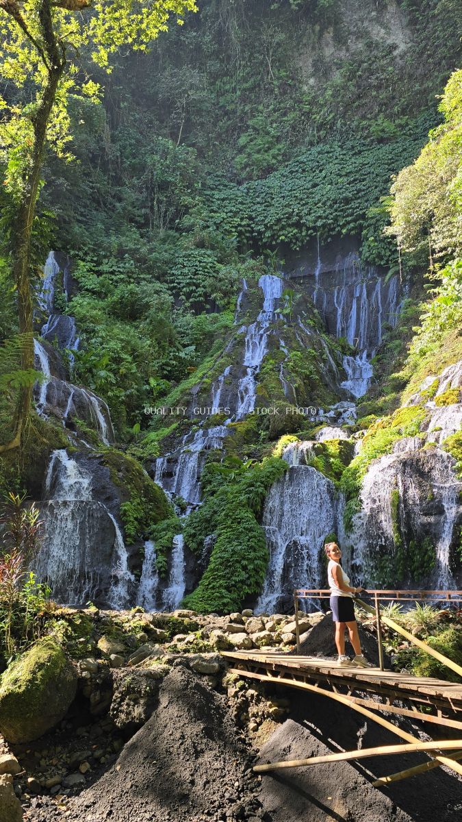 Woman hiker on wooden bridge viewing cascading tropical waterfall in Bali jungle with moss-covered rocks - stock photo