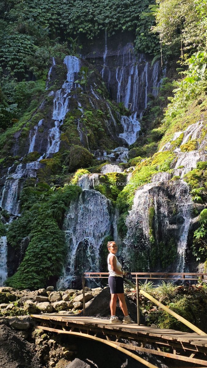 Traveler standing on wooden platform viewing cascading waterfall surrounded by lush green jungle vegetation in Bali - stock photo