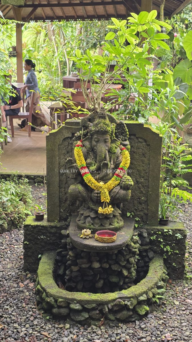 Moss-covered stone Ganesha statue with yellow flower garland in tropical Balinese garden pavilion - stock photo