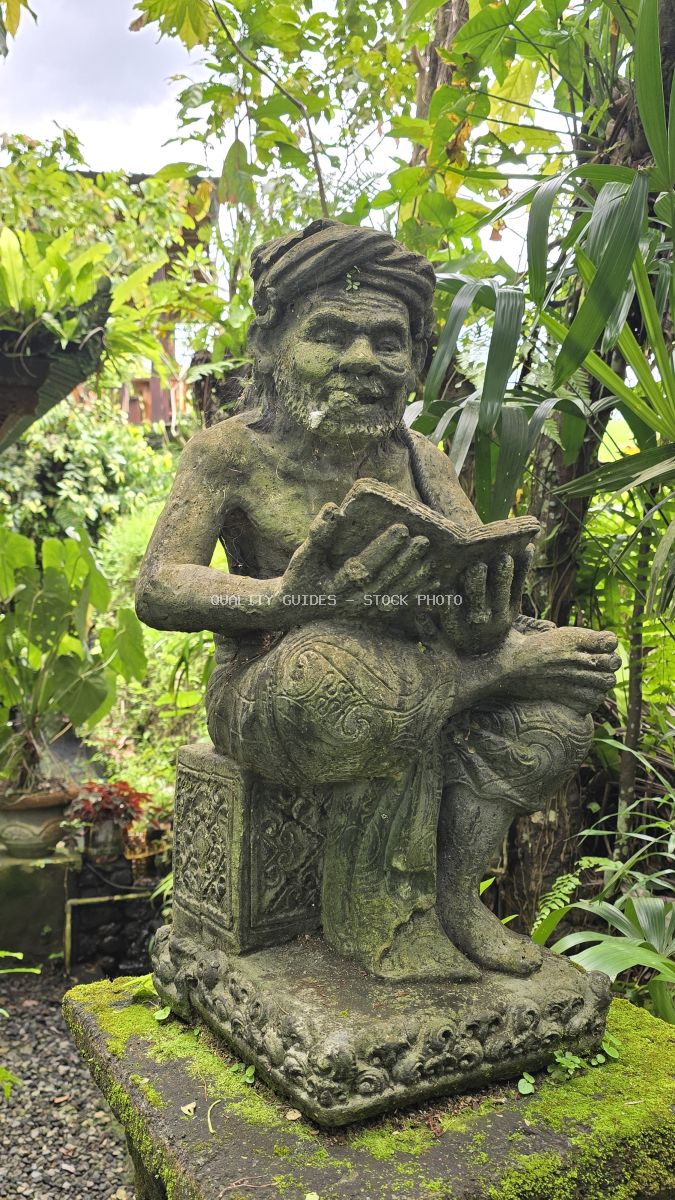 Weathered stone statue with bearded figure surrounded by tropical green foliage and moss in Bali garden - stock photo