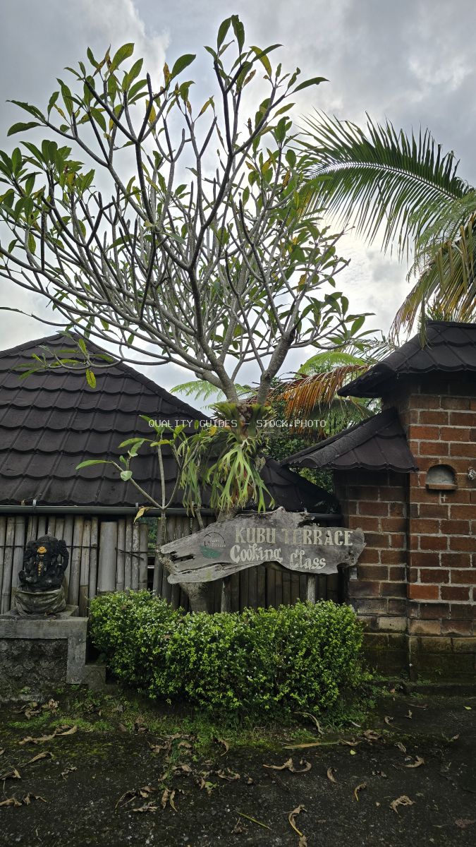 Balinese terrace restaurant with white plumeria tree, traditional dark-tiled architecture, and cooking class signage in Bali - stock photo