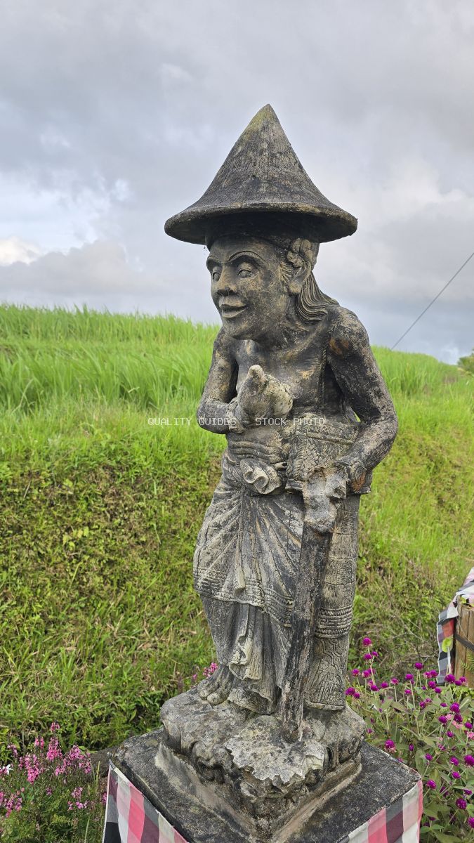 Weathered stone Balinese statue with conical hat overlooking green rice terraces at Jatiluwih, Bali - stock photo